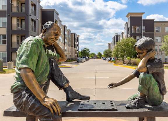 A statue of two people sitting on a bench in front of apartment buildings at Alinea Town and Country Luxury Apartments in Des Peres MO 63131..