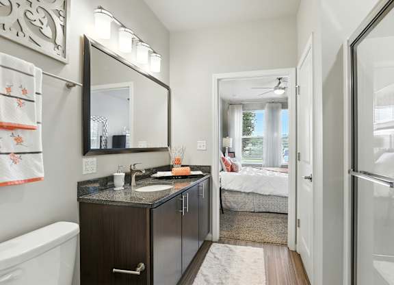 A bathroom with a sink, mirror, and a bedroom in the background at Alinea Town and Country Luxury Apartments in Des Peres MO 63131..