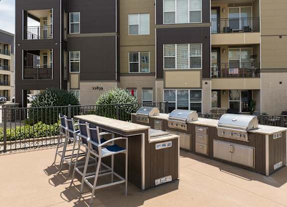 A patio with a grill and chairs is in front of apartment buildings at Alinea Town and Country Luxury Apartments in Des Peres MO 63131..