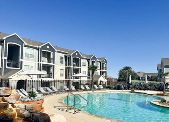 A swimming pool is surrounded by lounge chairs and umbrellas in front of apartment buildings.