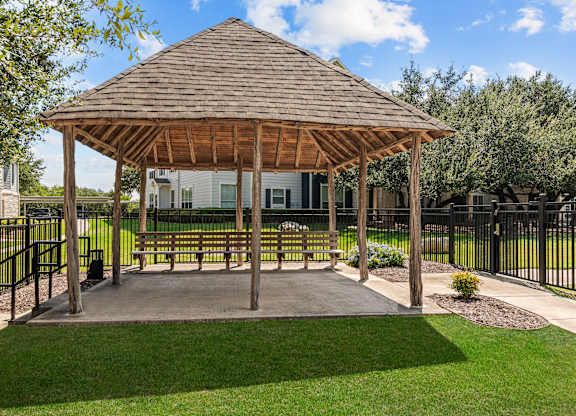A gazebo with a wooden roof is surrounded by a black fence and has a bench inside.