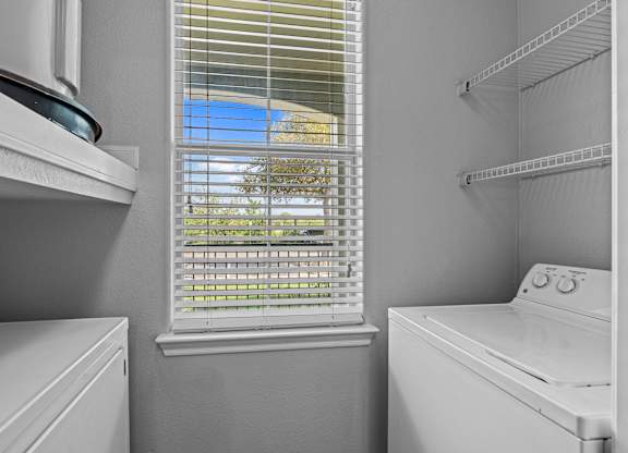 A white washing machine sits in a laundry room with a window covered in blinds.