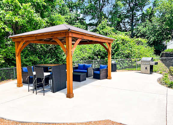 A wooden gazebo with a table and chairs is surrounded by trees.