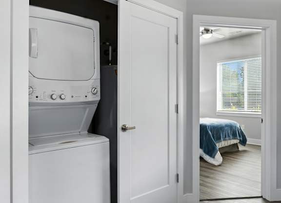 A white washing machine is in a small room with a bed in the background.