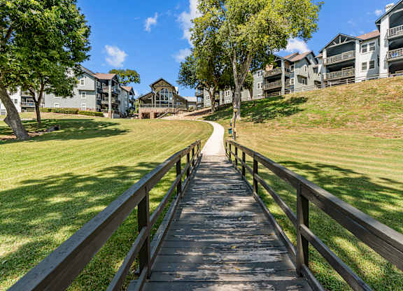 A wooden walkway leads through a grassy area with trees and apartment buildings in the background.