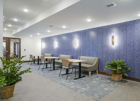 Cozy resident lounge seating area with small tables, upholstered chairs and bench seating, patterned accent wall, wall sconces, and indoor plants in a modern apartment community.