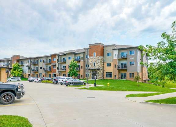 A modern stone and brok apartment building with grassy lawns, and a large parking lot.