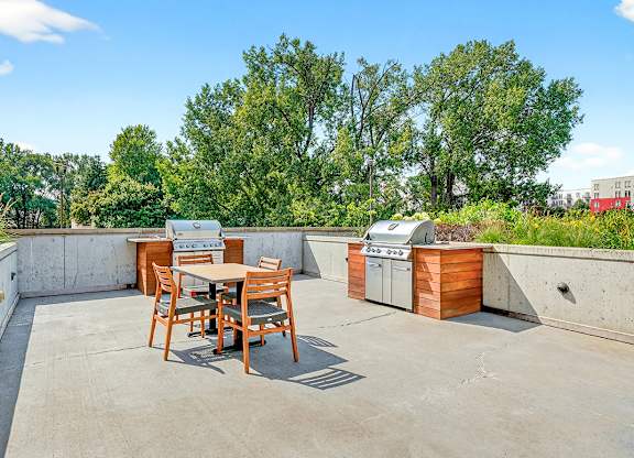 An outdoor grilling area with two built-in stainless steel grills, wooden cabinetry, and a dining table with chairs surrounded by lush greenery.
