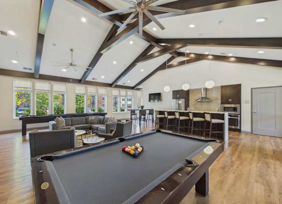 Apartment community game room with pool table, lounge seating, bar area, vaulted wood-beam ceilings, and large windows.