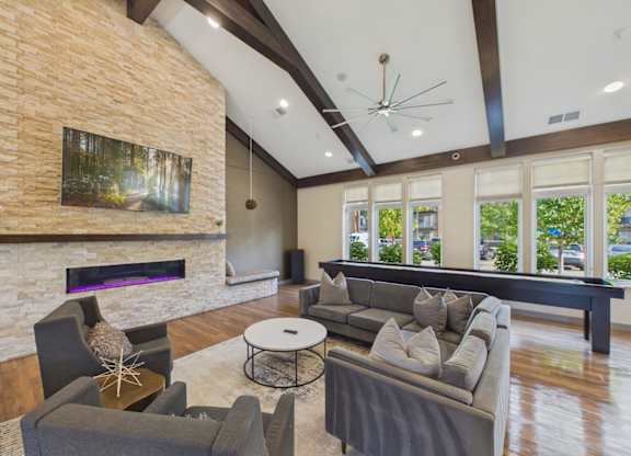 Bright apartment community clubhouse with vaulted wood-beam ceilings, stone fireplace, sectional seating, and large windows.