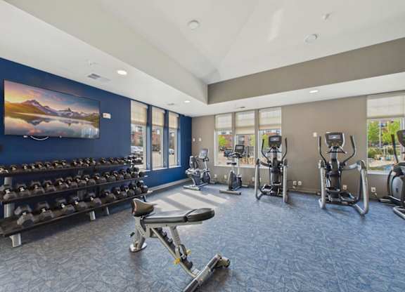 Apartment community fitness center with dumbbell racks, weight bench, elliptical machines, large windows, and blue accent wall.