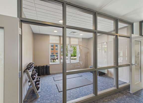 Apartment community fitness room with cardio equipment, rubber flooring, and glass walls overlooking the surrounding property.