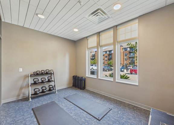 Apartment community fitness area with exercise mats, dumbbell rack, neutral walls, and large windows providing natural light.