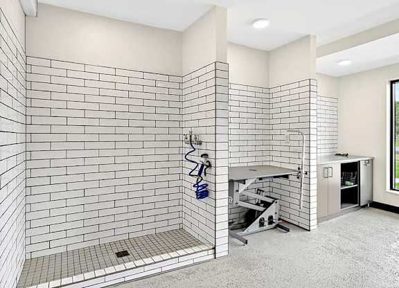 Indoor pet wash station with white subway tile walls, raised wash platform, handheld sprayer, and utility sink in a clean apartment amenity room.