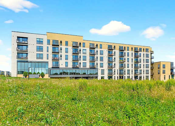 Modern apartment building exterior with multiple balconies, large windows, and mixed-tone facade overlooking open green space.