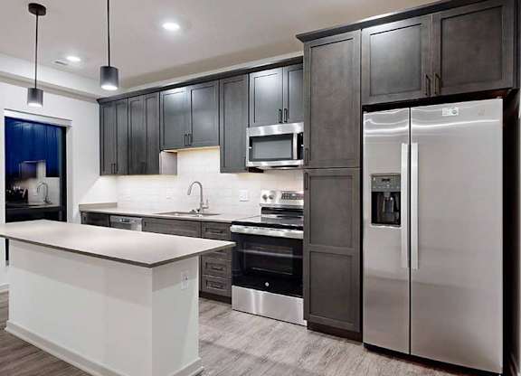 a modern kitchen with dark wood cabinets, light quartz countertops, a narrow central island, a tile backsplash, and stainless steel appliances