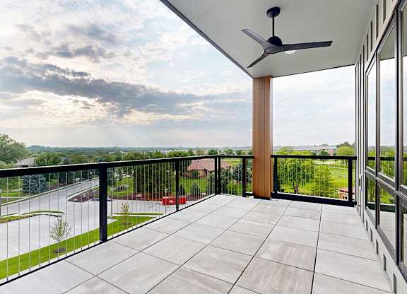 An oversized balcony with a metal and wire railing, stone tile flooring, a ceiling fan, and a corner view of the neighborhood.
