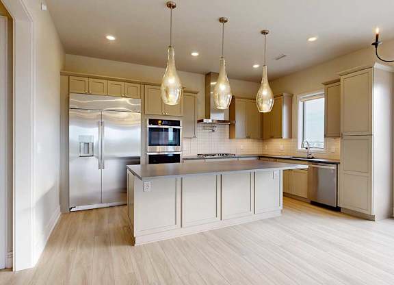 A gourmet kitchen with an oversized island, taupe wooden cabinetry, stainless steel commercial appliances, quartz countertops, a range hood, a tile backsplash, and pendant lighting.