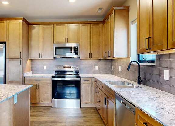 a kitchen with light brown wooden cabinets, stainless steel appliances, a tile backsplash, a large central island, and quartz countertops