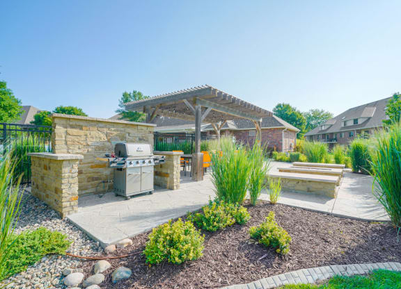a patio surrounded by greenery with a grill and a fire pit next to a pergola