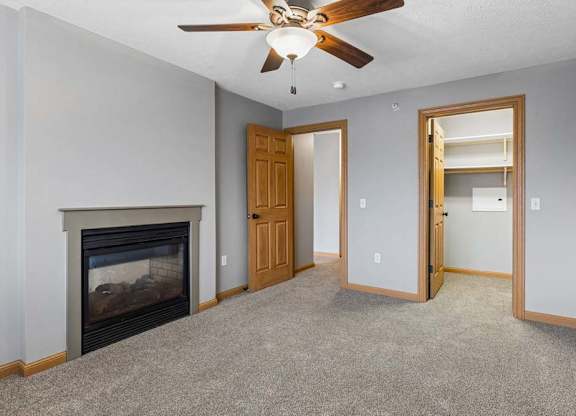 Apartment living room with neutral carpet, a corner fireplace, ceiling fan, and an open doorway leading to a laundry area with shelving.