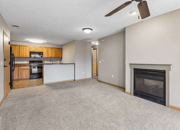 Open-concept apartment living room with carpet, a corner fireplace, ceiling fan, and breakfast bar overlooking a kitchen with wood cabinets and black appliances.
