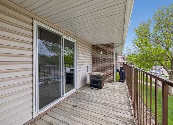 Private apartment balcony with wood decking, sliding glass door, metal railing, and views of mature trees and the surrounding community.