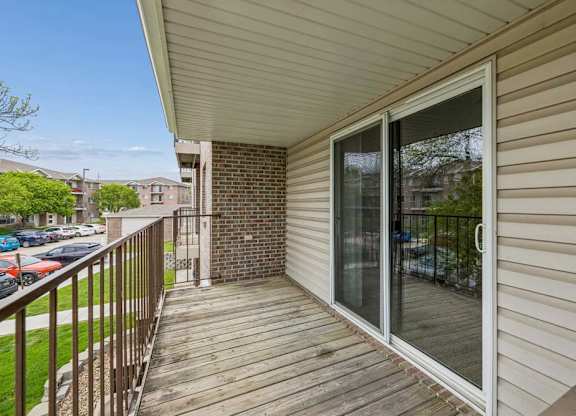 Spacious apartment balcony with wood decking, a sliding glass door, metal railing, and views of mature trees, nearby buildings, and community parking.
