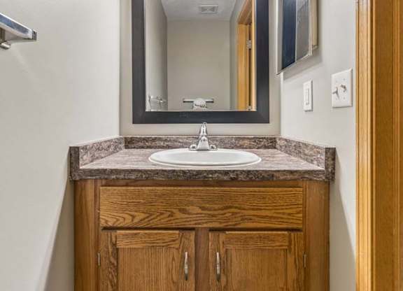 Compact apartment bathroom vanity with a wood-tone cabinet, laminate countertop, framed mirror, modern light fixture, and towel bar in a neutral interior.