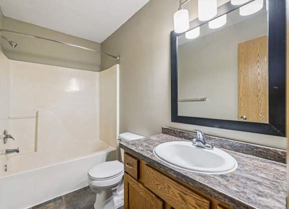 Apartment bathroom with a single-sink vanity, wood-tone cabinet, framed mirror, modern light fixture, toilet, and a tub/shower combination with built-in shelving.