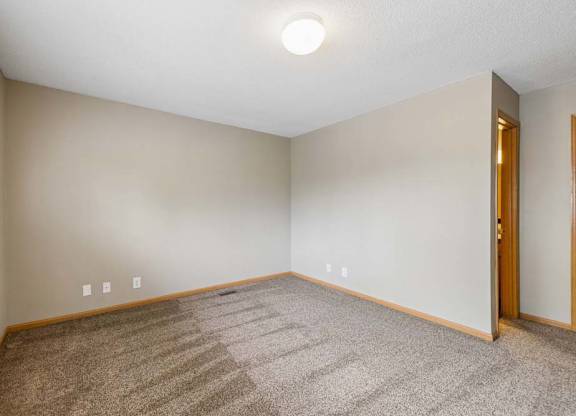 Empty apartment bedroom with neutral carpet, beige walls, wood trim, and an open doorway leading to the adjoining hallway.