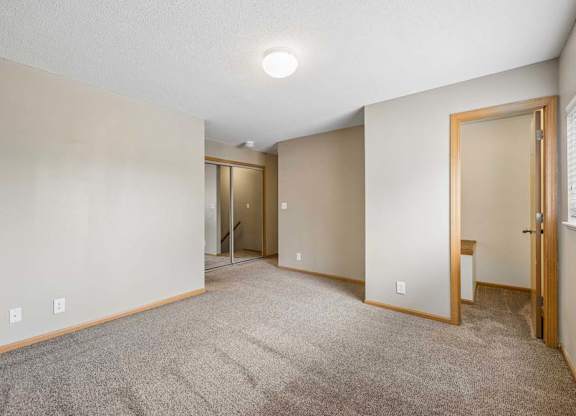 Bright, empty apartment bedroom with neutral carpet, beige walls, wood-trimmed doors, a window for natural light, and an open doorway leading to the closet area.