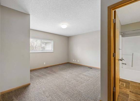 Bright apartment bedroom with neutral carpet, a window for natural light, and an adjoining bathroom with a single vanity and tub/shower visible through the open doorway.