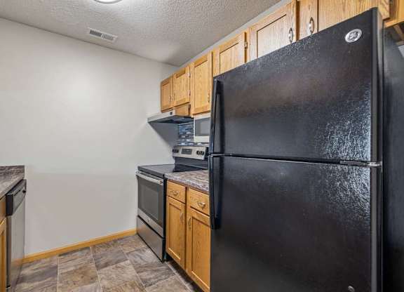 Galley-style apartment kitchen with wood-tone cabinets, black refrigerator and oven, laminate countertops, and durable flooring in a functional layout.