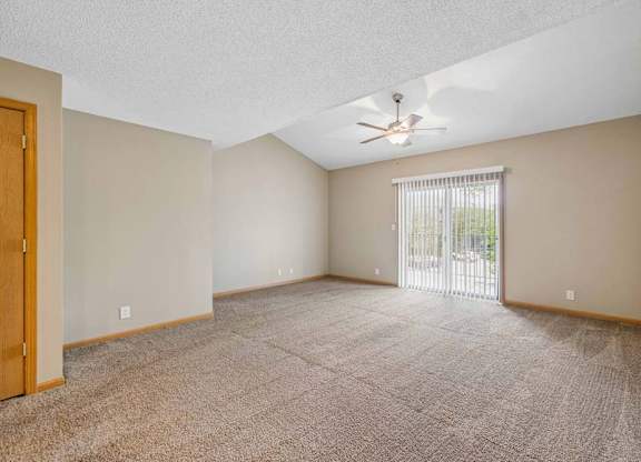 Spacious, empty apartment living room with vaulted ceilings, neutral carpet, a ceiling fan, and a large sliding glass door with vertical blinds that fills the space with natural light.