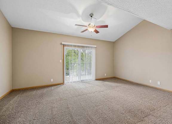 Bright, empty apartment living room with vaulted ceilings, neutral carpet, ceiling fan, and a large sliding glass door with vertical blinds that lets in natural light.