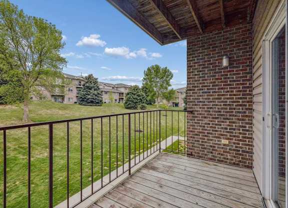 Covered apartment balcony with wood decking, a brick accent wall, metal railing, and views of the landscaped grounds and neighboring apartment buildings.