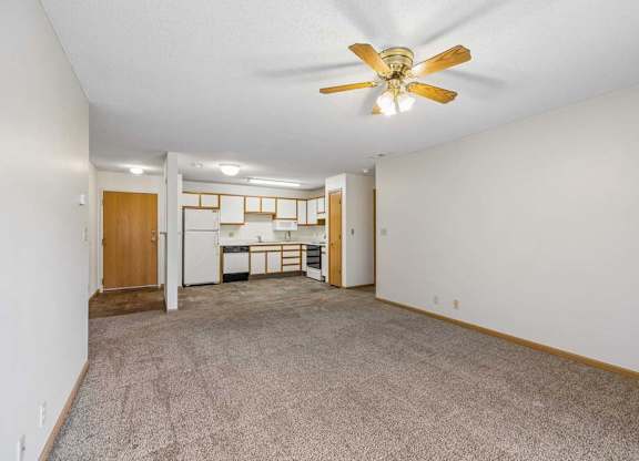 Spacious apartment living room with neutral carpet, a ceiling fan, and an open layout leading to the kitchen with a breakfast bar, wood-tone cabinets, and white appliances.