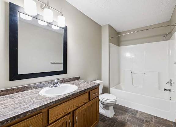 Apartment bathroom with a single-sink vanity, wood cabinetry, a granite-look countertop, a framed mirror with modern lighting, and a tub-shower combination with a white surround.