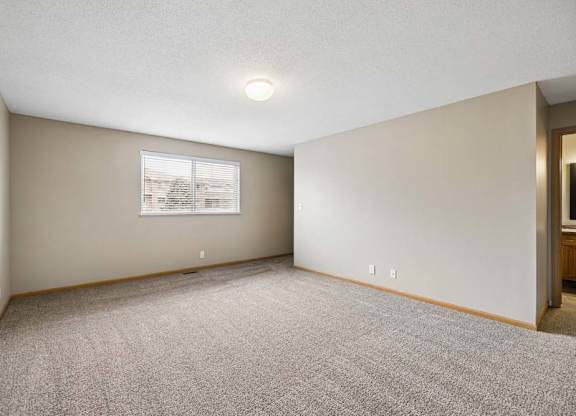 Spacious apartment bedroom with carpeted flooring, neutral walls, a large window providing natural light, and an open doorway leading to the adjoining bathroom.