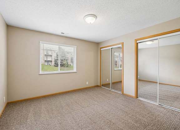 Bright apartment bedroom with carpeted flooring, a window providing natural light, and mirrored sliding closet doors for convenient storage.