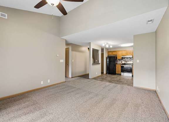 Open-concept apartment living room with carpeted flooring, a ceiling fan, vaulted ceilings, and a view into the kitchen featuring wood cabinetry and black appliances.