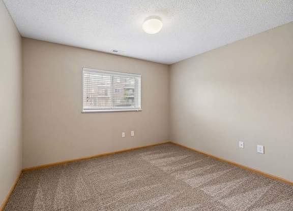 Bright apartment bedroom with carpeted flooring, neutral walls, and a window with blinds providing natural light.
