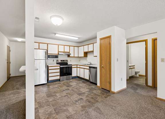 Open-concept apartment with a kitchen featuring white and wood-tone cabinetry, stainless steel and black appliances, a pantry, and adjacent carpeted living and dining areas.