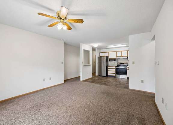 Spacious open-concept apartment living room with carpeted flooring, a ceiling fan, and a view into the kitchen featuring white and wood-tone cabinetry, stainless steel and black appliances, and a functional open layout.