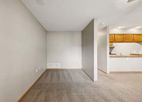 Spacious apartment living area with carpeted flooring and a flexible nook, adjacent to an open kitchen with a breakfast bar and wood cabinetry.