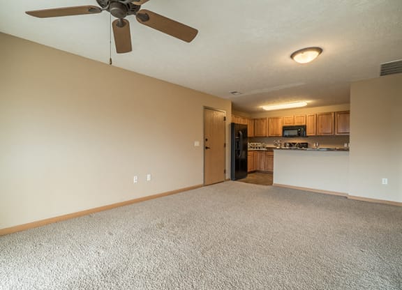 a spacious living room with a ceiling fan, next to a kitchen with wooden cabinets and a tall bar top counter