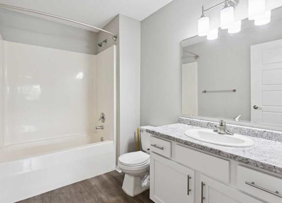 Close-up of bathroom vanity with white cabinetry, speckled countertop, oval sink, wide mirror, and modern three-light fixture above, with toilet beside.
