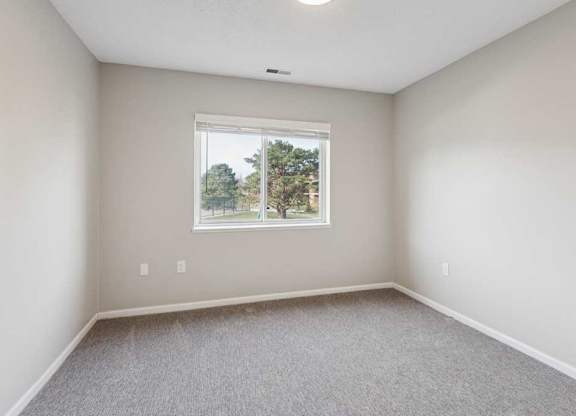 Bright bedroom with carpet flooring and a wide window overlooking neighboring buildings, providing natural light and a clean, neutral interior.