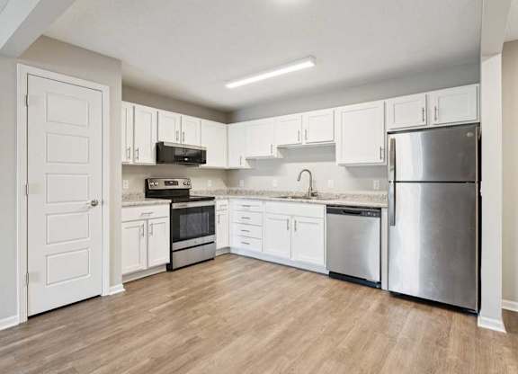 Modern apartment kitchen with white cabinets, speckled countertops, stainless steel refrigerator, oven, microwave, and dishwasher, plus wood-style flooring.